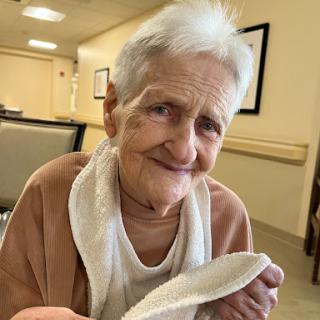 An elderly woman smiles warmly while seated indoors, holding a towel.
