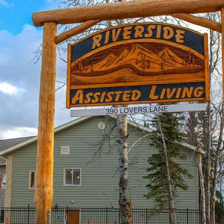 A wooden sign for Riverside Assisted Living stands in front of a building surrounded by trees.