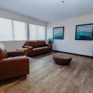 A modern living room featuring brown leather furniture, a round coffee table, and wall art, illuminated by natural light from large windows.