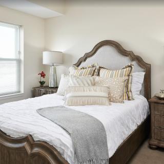 A cozy and elegantly decorated bedroom with a large bed, decorative pillows, a lamp, and a window allowing natural light.