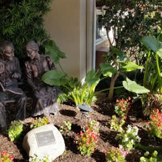 A serene garden scene featuring a sculpture of two figures engaged in conversation amidst vibrant plants.