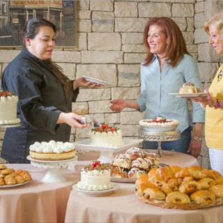 A chef discusses a variety of desserts with two women at a bakery setup.