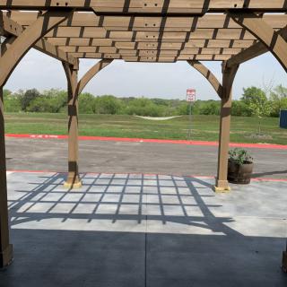 A wooden pergola casting shadows on a sunny day, overlooking a grassy landscape.