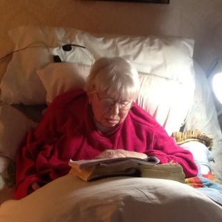 An elderly person reading a book in bed with a lamp providing light.
