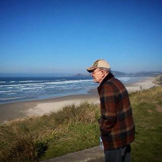 A man wearing a cap and jacket stands by the coastline, gazing at the ocean under a clear blue sky.