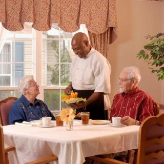 A waiter serves a meal to two seniors at a dining table indoors.