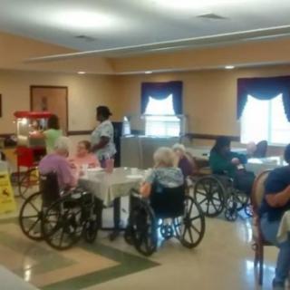 Elderly residents in wheelchairs gathered in a common area of a nursing home with staff members nearby.
