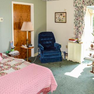 A cozy bedroom with vintage decor, featuring a bed, armchair, lamp, and window with floral curtains.
