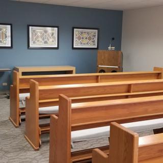 A small chapel interior featuring wooden pews and framed artworks on the wall.