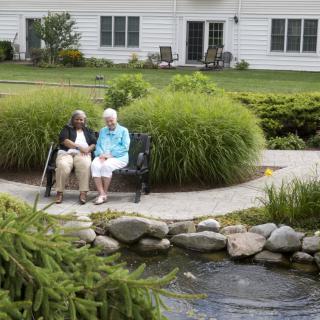 An elderly couple sits on a bench enjoying the serene garden and pond view.