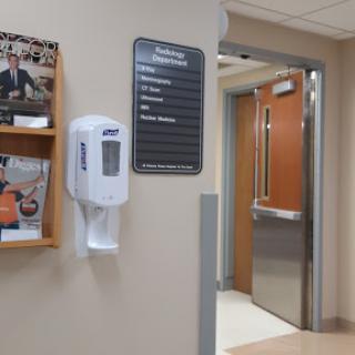 A view of a hospital waiting area featuring a sanitizer dispenser, an information sign, and magazines.