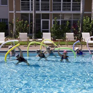 A group of people exercising with pool noodles in an outdoor swimming pool.