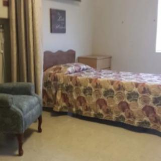 A cozy bedroom featuring a single bed with a floral bedspread, an armchair, and light-colored curtains.