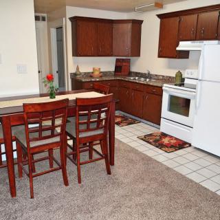 A cozy kitchen with wooden cabinets and a dining area, featuring white appliances and a carpeted floor.