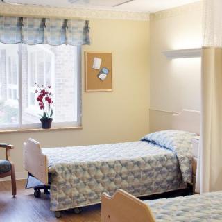 A clean and well-lit hospital room with two beds, featuring a window with curtains and a plant on the windowsill.
