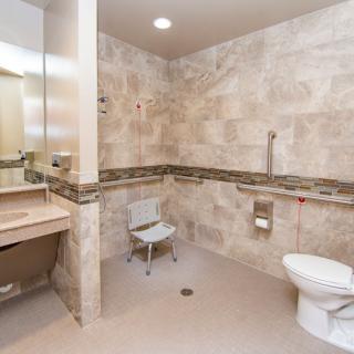 An accessible bathroom featuring a toilet, sink, and safety chair with grab bars installed on the tiled walls.