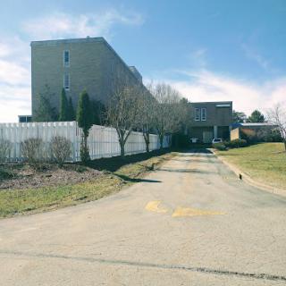 An abandoned building along a deserted road, surrounded by a fence and sparse vegetation.