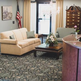 A cozy waiting room with a sofa, armchair, and coffee table adorned with a floral arrangement. An American flag stands in the corner, and the space features elegant curtains and a detailed carpet.