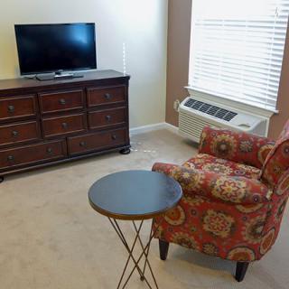 A cozy living room corner with a patterned chair, side table, dresser, and a TV near a window.