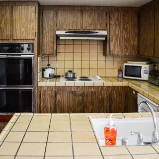 A retro-style kitchen with wooden cabinets and tiled countertops, featuring a microwave, oven, and sink.