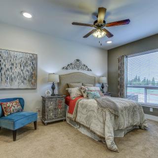 A cozy and elegantly furnished bedroom featuring a large bed with decorative pillows, a patterned rug, artwork on the wall, a blue armchair, and a ceiling fan.