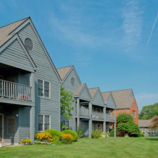 A row of modern apartment buildings with balconies, set against a clear blue sky and surrounded by neatly maintained green grass and landscaping.
