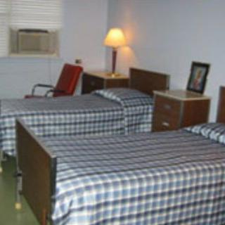 A simple bedroom with twin beds featuring checkered sheets, an air conditioner in the window, a lamp on a table, and a chair.