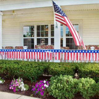 A porch decorated with American flag-themed bunting and an American flag, surrounded by a garden with colorful flowers.