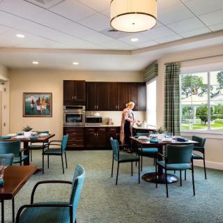 A modern dining room with a kitchen area, featuring tables and chairs, where a woman is setting up the space.