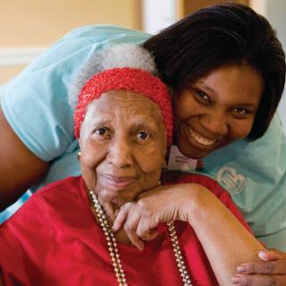A smiling caregiver embraces an elderly woman wearing a red headband and shirt.