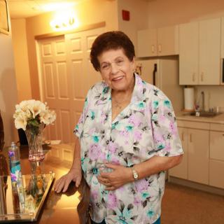 An elderly person smiling in a kitchen area, wearing a floral shirt with flowers on the counter.