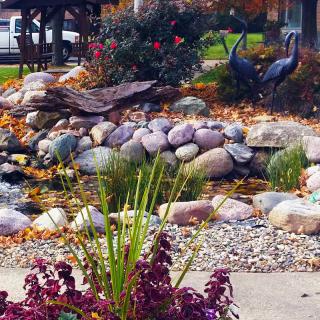 A beautifully landscaped garden featuring a small pond surrounded by rocks and plants, with sculptures enhancing the scene.