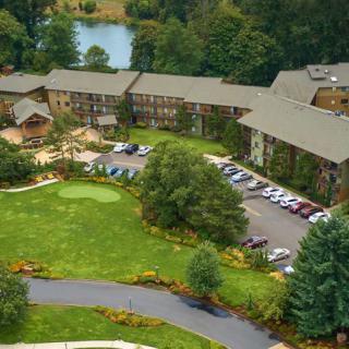 Aerial view of a building complex surrounded by lush greenery and trees, featuring a neatly maintained lawn and parking area.