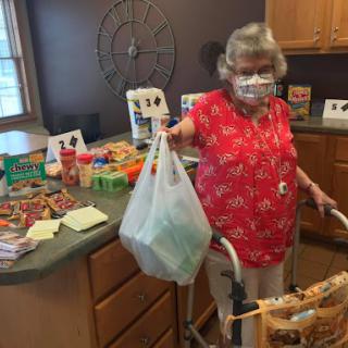 An elderly woman in a red blouse is holding a bag of groceries while using a walker in her kitchen.