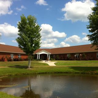 A serene view of a building with a pond in the foreground and a clear sky with scattered clouds.