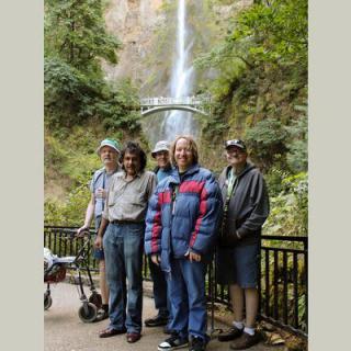 A group of people standing in front of a waterfall and a bridge, enjoying the scenic outdoor setting.