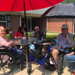 A group of senior citizens enjoying a sunny day outside, gathered around a table under a large red umbrella.