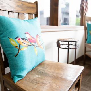 A wooden chair with a colorful bird-themed pillow on a bright indoor patio.