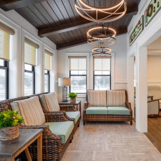 A cozy and well-lit living room featuring wicker furniture, modern chandeliers, and large windows that bring in natural light.