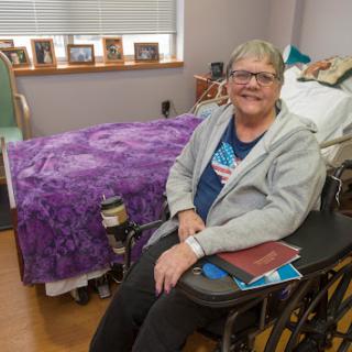 A smiling individual seated in a wheelchair, surrounded by personal belongings in a cozy room.
