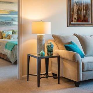 A cozy living room with a sofa, side table, and lamp, leading into a well-decorated bedroom.