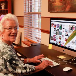 An elderly woman smiling while using a computer at her desk in an office setting.