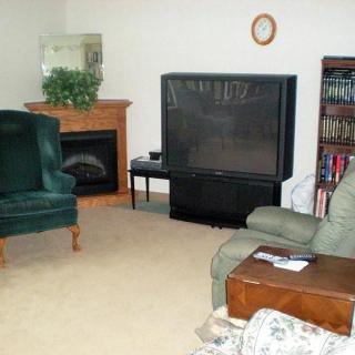 A cozy living room with a large television, armchairs, a fireplace, and a bookshelf.