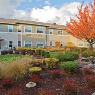 A landscaped garden with shrubs and a tree in autumn colors, situated in front of a two-story building under a cloudy sky.
