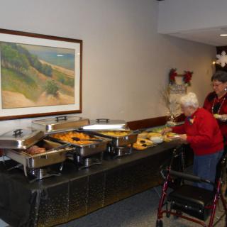 Elderly woman with a walker selecting food from a buffet in an indoor setting.