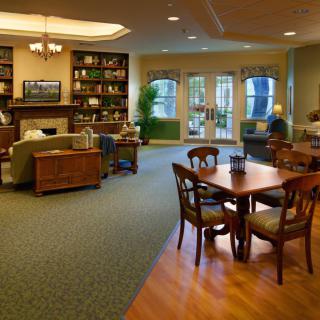A cozy living room featuring a sofa, bookshelf, wooden table with chairs, and large windows.