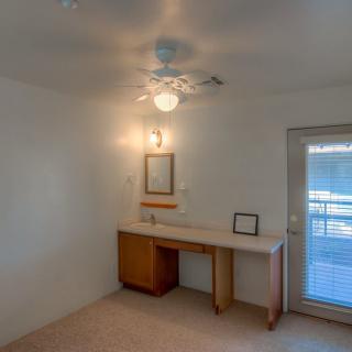 A minimalistic room with a small desk, mirror, ceiling fan, and carpeted floor.