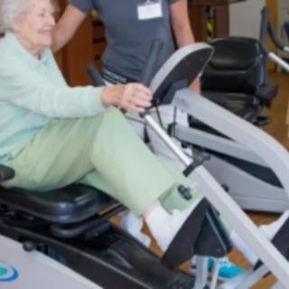 An elderly woman is exercising on a recumbent bike with assistance from a caregiver.