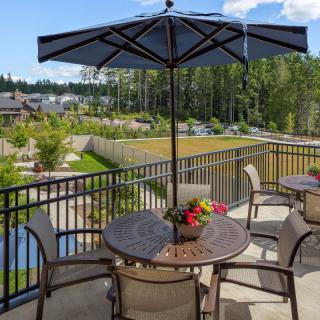 A serene patio with a round table and chairs under an umbrella, overlooking a lush garden and distant water body.