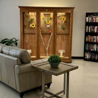 A cozy lounge area featuring a leather couch, a wooden cabinet with sunflowers, and a bookshelf filled with books.